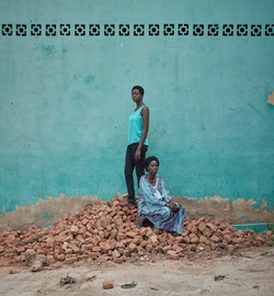 Zwei Frauen der Rwandan Daughters vor einer türkisfarbenen Hauswand mit Verzierung. Eine der Frauen steht, die andere sitzt neben ihr auf einem Berg mit rötlichen Ziegelsteinen. 